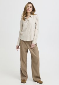 Mujer de pie con una camisa beige abotonada, pantalones sueltos color arena y zapatos marrones, frente a un fondo claro y sencillo.