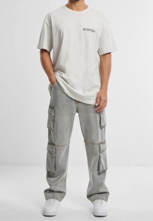 Mann trägt hellgraues übergroßes T-Shirt und graue Cargo-Jeans mit mehreren Taschen, kombiniert mit weißen Turnschuhen, steht mit einer Hand in der Tasche.