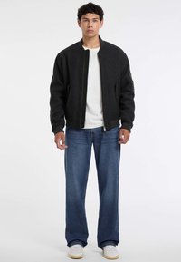 Giacca bomber nera con polsini aderenti, chiusura con zip, tasche laterali, indossata sopra una camicia bianca. Abbinata a jeans blu larghi e sneakers bianche.