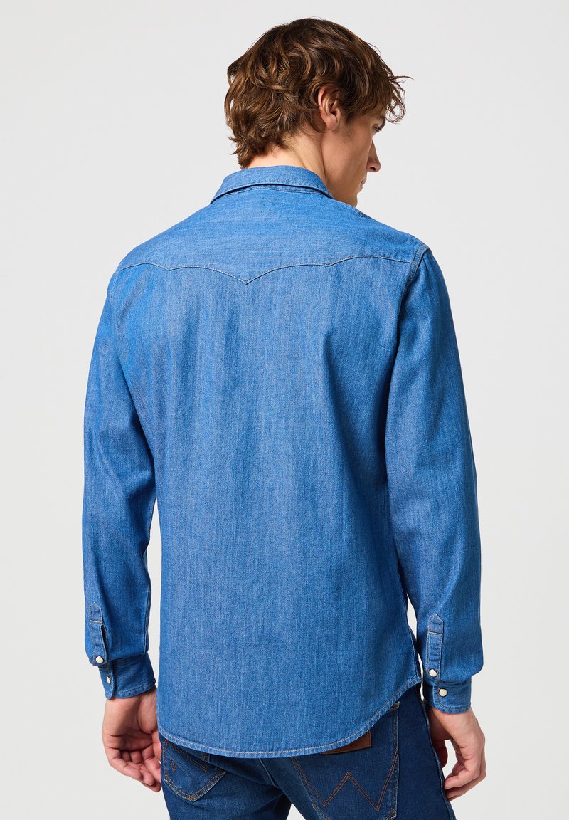 Camisa de mezclilla en azul medio, de manga larga, con cuello puntiagudo, y yugo trasero con un discreto detalle de costura, y puños con botón. Textura suave.
