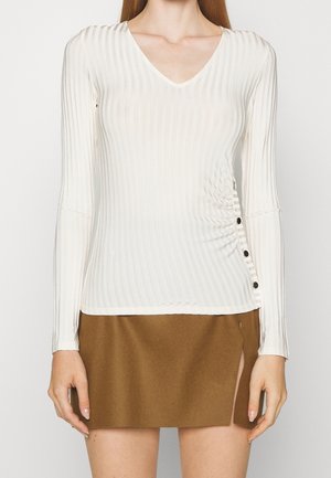 Haut blanc côtelé à manches longues avec détail de boutons sur le côté, associé à une mini-jupe marron avec une fente à l'avant.