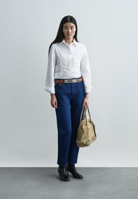 Camisa blanca ajustada con detalles fruncidos, jeans azules de cintura alta, botines negros y bolso marrón claro. Cinturón con el logo "MOSCHINO".