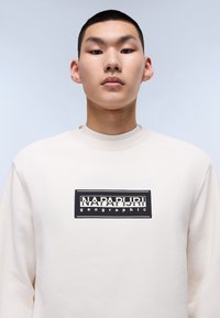 Felpa color crema con scollatura rotonda, caratterizzata da un logo rettangolare nero e testo sul davanti. Tessuto liscio e vestibilità casual.