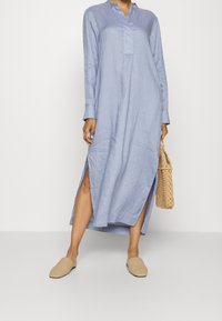 Kobieta w długiej, jasnoniebieskiej sukience z bocznymi rozcięciami, trzymająca plecioną torbę i beżowe, plecione pantofle wsuwane.