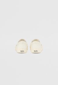 Pantofi slip-on din canvas bej, cu vârf rotund, talpă plată și un detaliu cu logo negru pe călcâi. Design minimalist, fără modele.