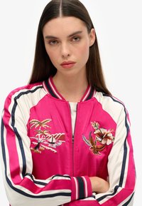 Veste bomber en satin rose avec des manches blanches, ornée de broderies florales colorées et d'accents rayés. Poignets et col côtelés.
