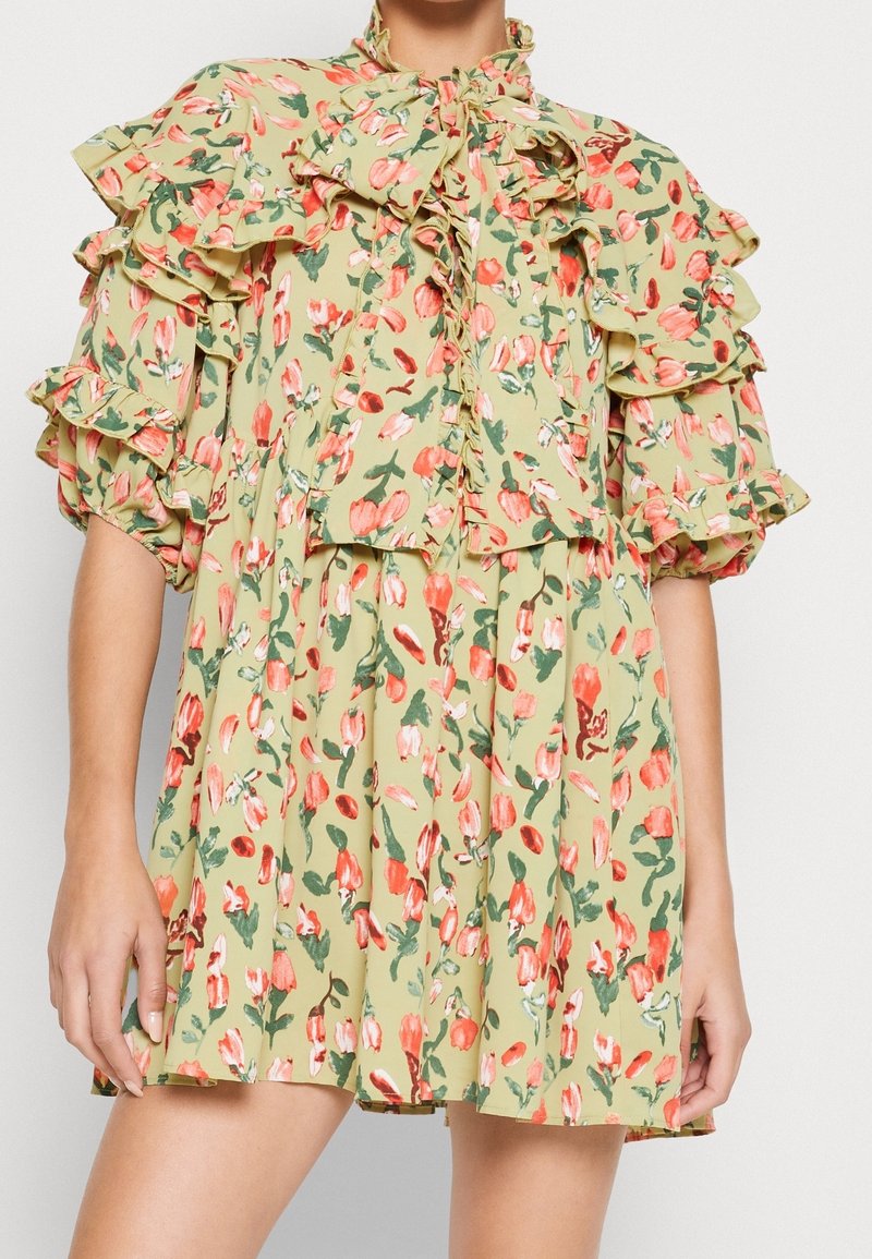 Hellgrünes Blumenkleid mit gepufften Rüschensäulen, einer großen Schleife am Hals und einem gefältelten Rock, getragen von einer stehenden Person.