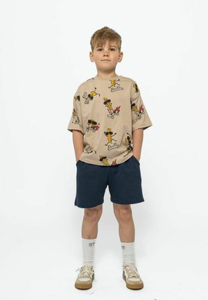 Camisa beige de manga corta con divertidas ilustraciones de patinetas, combinada con pantalones cortos azul marino. Zapatillas ligeras y calcetines blancos completan el conjunto.