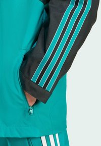 Turkos jacka med svarta ärmar, med tre vita ränder längs armarna och en dragkedjeficka på sidan. Slät textur.