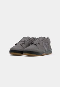 Grå semsket lær sneakers med lavt snitt, svart gummisåle, snøring foran og en subtil logodetalj på siden.