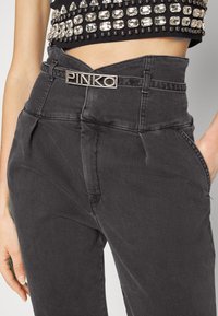 Pantalones vaqueros negros de cintura alta con un ligero brillo, que cuentan con bolsillos laterales y un logo metálico "PINKO" en la cinturilla.