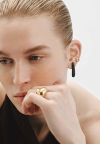 Pendientes en dorado y negro, con un aro negro texturizado y un arete redondo en dorado. También es visible un anillo dorado escultórico y brillante.