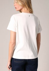 T-shirt en coton blanc à manches courtes, coupe décontractée, avec un petit logo sur le dos. Porté avec un jean bleu foncé.