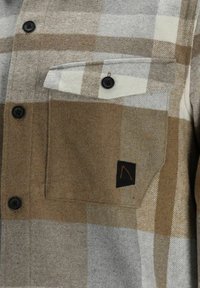 Camisa a cuadros en tonos marrón y gris con un bolsillo en el pecho, botones negros y una etiqueta negra de la marca. Fabricada con un tejido suave y texturizado.
