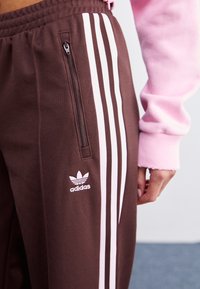 Hnědé sportovní kalhoty s bílým logem Adidas a třemi svislými bílými pruhy po stranách; včetně kapsy na zip na levé noze.