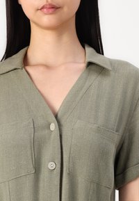 Chemise à manches courtes, verte claire, en tissu texturé, avec un col, deux poches avant et une fermeture à boutons.