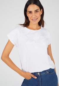 Witte T-shirt met korte mouwen, featuring bloemenborduursel op de voorkant, gemaakt van zachte stof. Gecombineerd met donkere spijkerbroek.