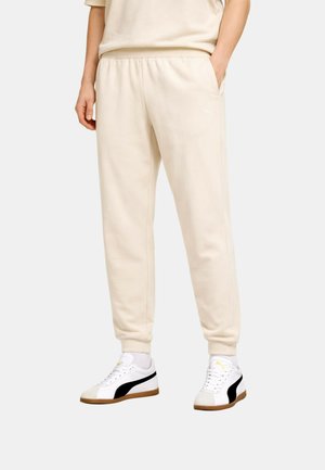 Person, die beige Jogginghosen trägt, die Hände in den Taschen hat und weiße Turnschuhe mit schwarzen und gumfarbigen Sohlen trägt, steht vor einem einfarbigen Hintergrund.