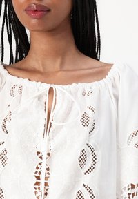 Weiße bestickte Bluse mit rundem Ausschnitt, gerafftem Stoff und Bindeschluss. Verfügt über aufwendige Muster und durchsichtige Abschnitte.
