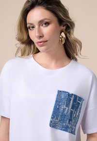 Camiseta blanca de manga corta con un parche de denim azul adornado con lentejuelas. Ajuste relajado, con escote redondo y textura suave.