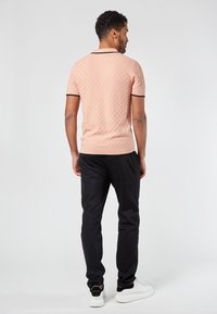 Persikkavärinen teksturoidulla pinnalla oleva polo-paita, jossa on lyhyet hihat ja musta reunus kauluksessa ja hihoissa, yhdistetty mustiin kapeisiin housuihin ja valkoisiin lenkkareihin.