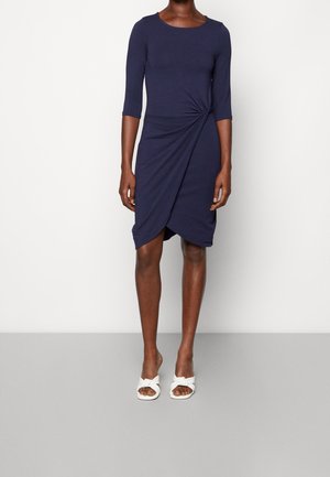 Femme portant une robe bleu marine longueur genoux avec des manches trois-quarts et un détail de nœud torsadé à la taille, associée à des talons ouverts blancs.