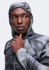 Homme portant une veste à capuche grise avec une fermeture éclair, regardant sur le côté la main touchant son menton.