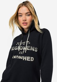 Sudadera negra con capucha y cordón, y bolsillo tipo canguro en la parte frontal. Presenta texto blanco en negrita y patrones gráficos en el pecho. Textura de tela suave.