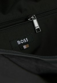 Intérieur d'un sac en tissu noir avec une fermeture éclair argentée et une étiquette portant "BOSS" en lettres blanches. Des accents incluent des coutures multicolores.