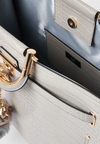 Lys grå handbag i krokodilleprægning med guldfarvet hardware, der har et rummeligt interiør, en tryklås lomme og en pels pom-pom nøglering som detalje.