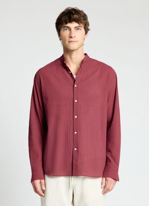 Jeune homme portant une chemise bordeaux texturée à manches longues avec des boutons blancs et un pantalon clair sur un fond uni.