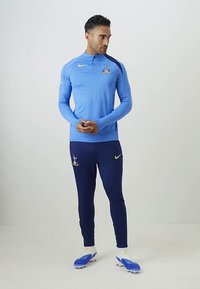 Zils treniņkrekls ar augstu apkakli un izšūtu logo, apvienots ar tumši ziliem biksēm, kas arī ir ar logo un kontrastējošām dzeltenām detaļām; uzvilkti futbola krekli.