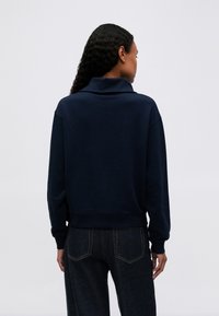 Femeie cu păr creț, îmbrăcată cu un top de culoare bleu marin cu mâneci lungi și guler înalt, purtând blugi negri, întorsă cu spatele, pe un fundal simplu.