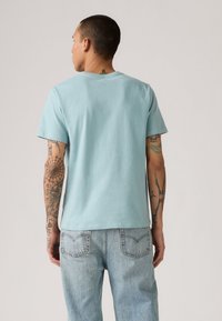 Tricou din bumbac de culoare bleu deschis, cu mâneci scurte, guler rotund și o croială lejeră. Poartă blugi deschiși, evidențiind tatuaje pe spate.