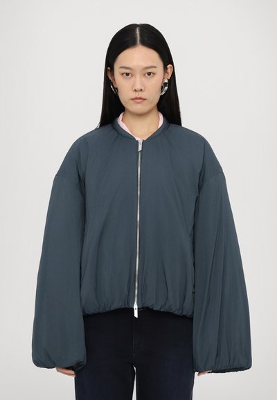 Veste bomber matelassée en gris foncé avec un col haut, fermeture éclair à l'avant et des manches volumineuses. Texture lisse et détail d'ourlet élastiqué.