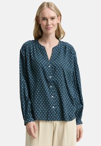 Blouse à manches longues en bleu sarcelle foncé avec un motif géométrique. Dispose d'un col en V, d'une fermeture à boutons et d'une coupe décontractée avec des épaules froncées.