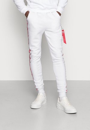 Person trägt weiße Jogginghosen mit roten Seitenstreifen und weiße Sneakers, Hände in den Taschen, steht vor einem einfarbigen Hintergrund.