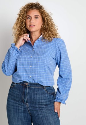 Femme aux cheveux blonds bouclés portant une blouse à boutons à carreaux bleus à manches longues et un jean bleu foncé taille haute.