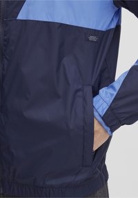 Veste coupe-vent bleu marine et bleu clair, dotée d'une fermeture éclair, d'une poche latérale et de poignets élastiques, fabriquée en tissu lisse et léger.