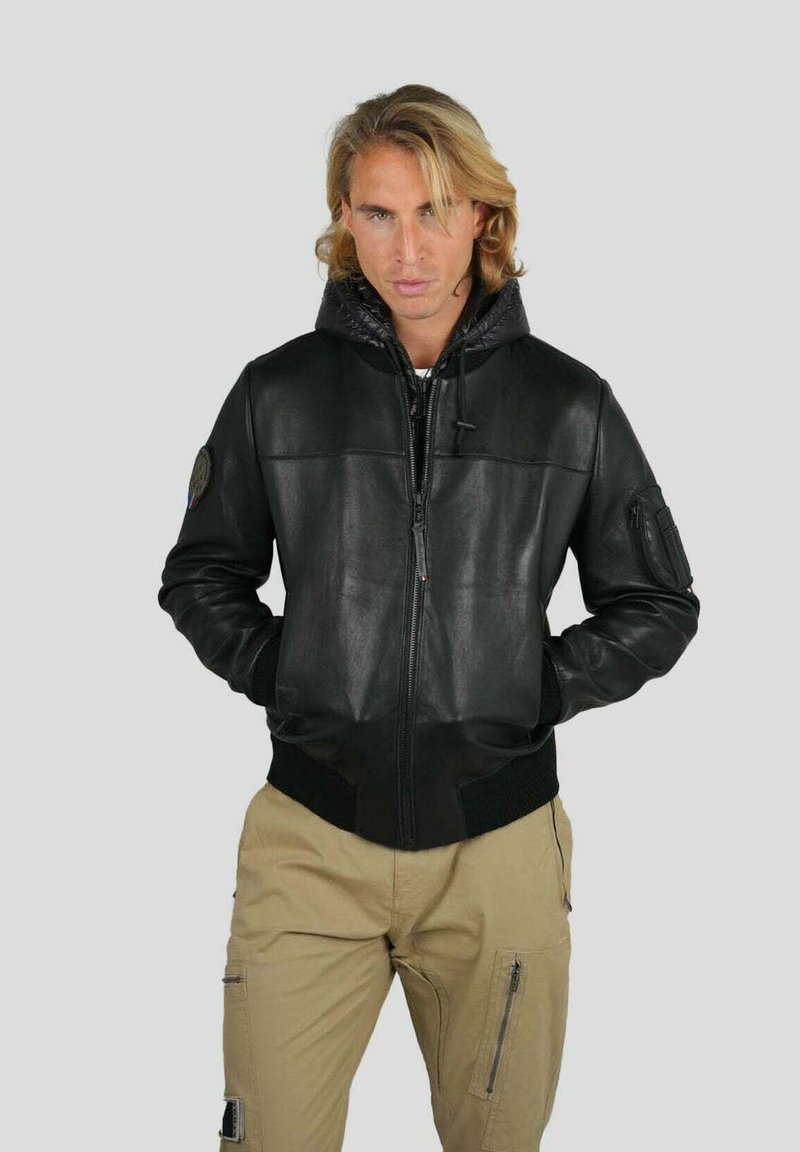 Homme blond avec les mains dans les poches portant une veste en cuir noir avec fermeture éclair et capuche, associée à un pantalon cargo beige sur fond uni.