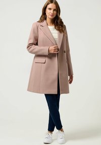 Rozige wollen blend jas met een gekante kragen, twee zijzakken, knoopsluiting en een relaxte pasvorm, gecombineerd met marineblauwe broeken en witte sneakers.