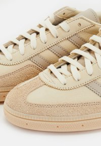 Beige tennarit, joissa on mokkanahka- ja nahkapäällinen, pyöreä kärki, litteät beige nauhat ja teksturoitu kumipohja, jossa on geometrinen kuvio.