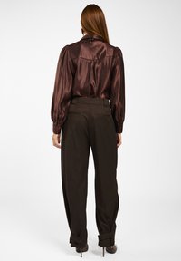Bruine satin blouse met geplooide mouwen en een rugpand, gecombineerd met donkerbruine op maat gemaakte broeken met knoopdetails aan de manchetten.