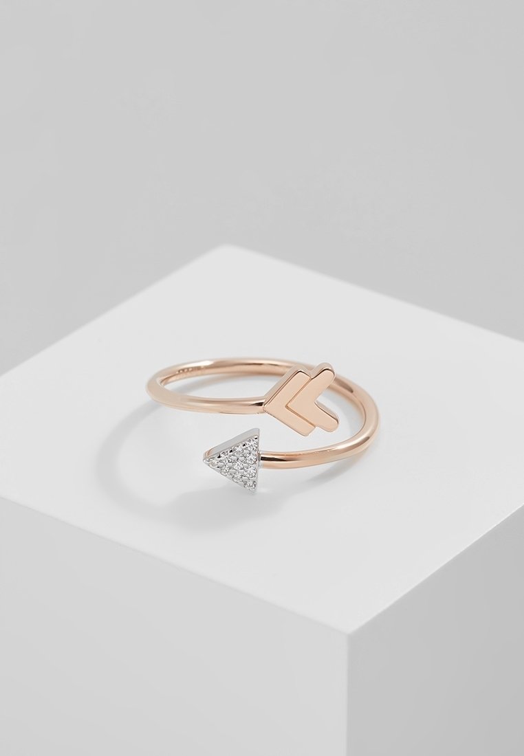 Bague ouverte en or rose avec un design en forme de pointe de flèche, une extrémité lisse et l'autre ornée de petits cristaux clairs, présentée sur une plateforme blanche.