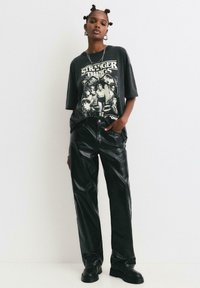 Crna majica s grafikom koja prikazuje "Stranger Things", u kombinaciji s sjajnim crnim lašnim kožnim hlačama i crnim gležnjačama. Opusteni kroj.