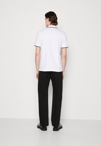 Weißes Poloshirt mit schwarzem Saum, kombiniert mit schwarzen Hosen. Der Stoff wirkt glatt, und das Hemd hat ein klassisches Kragendesign.