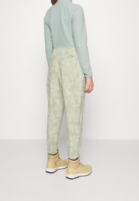 Ljust gröna kamouflagemönstrade cargobyxor med elastiska muddar och dragsko i midjan, tillsammans med bruna vandringskängor, sedd bakifrån.