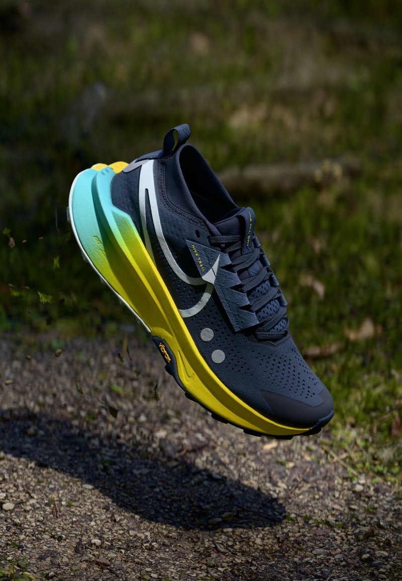 Chaussure de course bleu foncé et jaune flottant au-dessus d'un chemin de gravier avec un arrière-plan naturel vert flou.