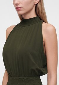 Femme portant une robe sans manches vert foncé avec un col montant froncé et une taille smockée, montrée de l'épaule au menton.