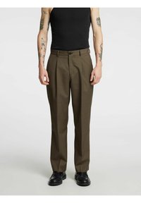 Bruine geplooide broek met een getailleerde pasvorm, gemaakt van een gestructureerde stof. Voorzijde met knoopsluiting en twee zijzakken. Gecombineerd met zwarte schoenen.